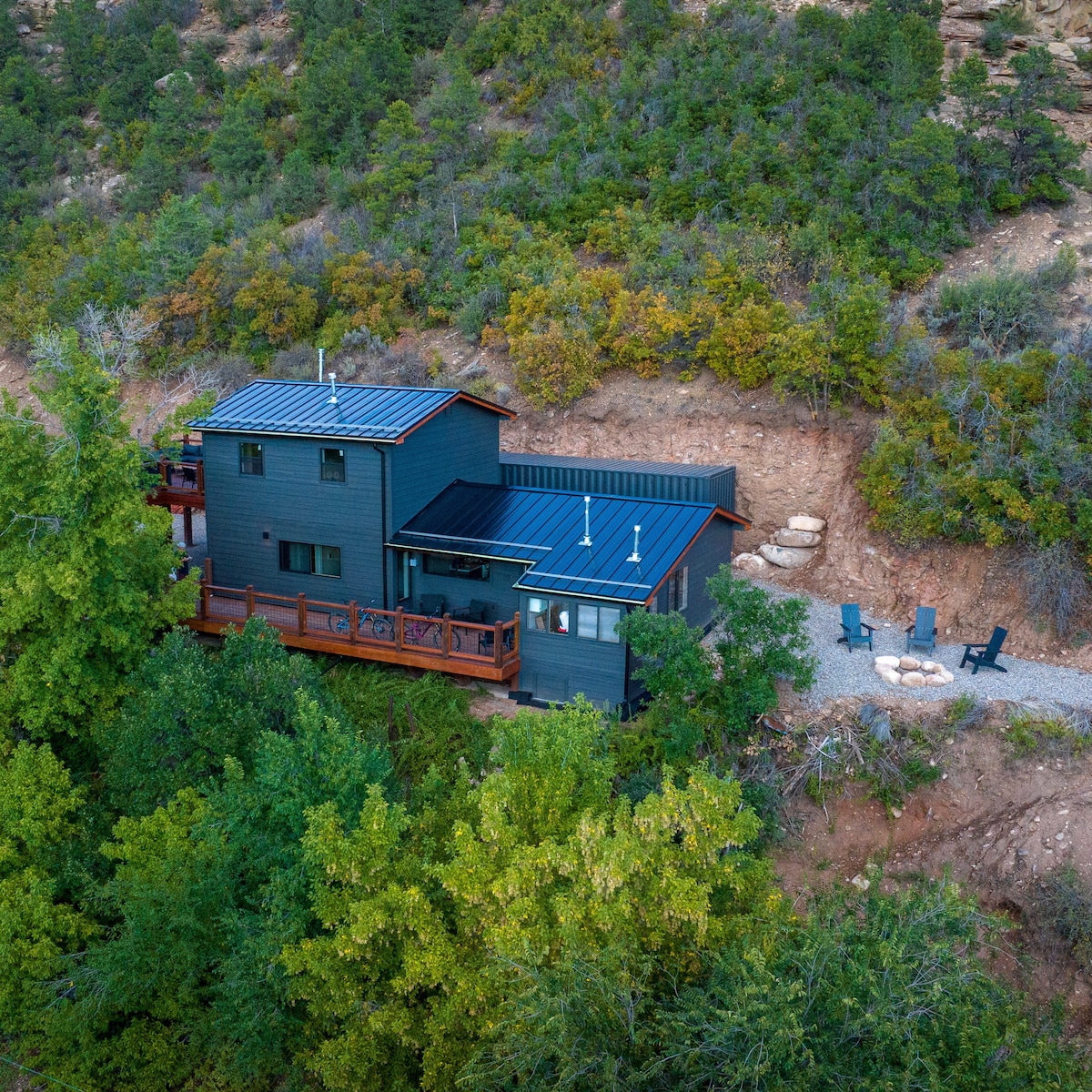 The exterior of Casa de Sendero is set against a backdrop of lush greenery, featuring a dark-colored home with a modern design. A spacious deck surrounds the building, complemented by an outdoor seating area with fire pit and chairs.