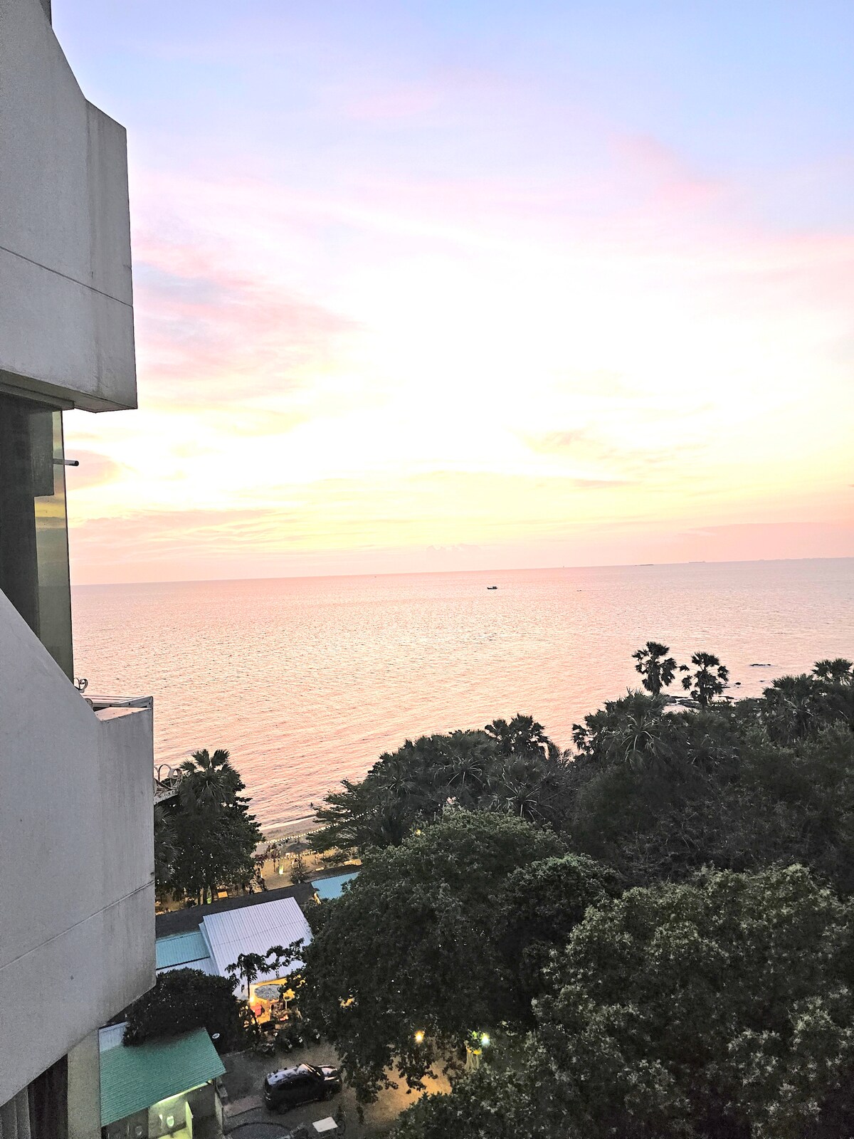 A panoramic view of the ocean is captured during sunset, showcasing soft pastel colors across the sky. The reflection on the water adds a calming ambiance. Tropical trees and buildings are visible in the foreground, enhancing the scenic coastal setting.