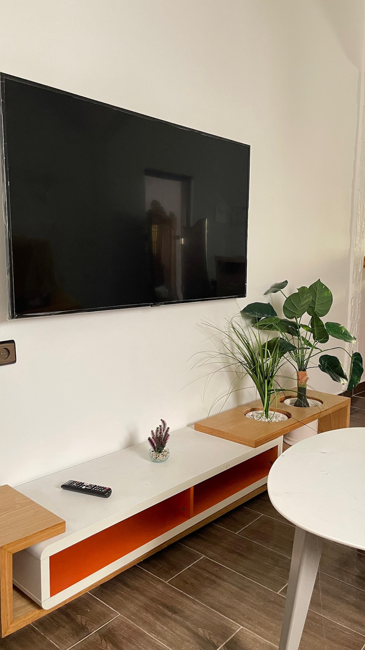 A wall-mounted flat-screen television is positioned above a light wooden media console. The console features an orange accent panel and is complemented by decorative plants. A remote control rests on the surface, with a small potted succulent and grass arrangement adding a touch of greenery.