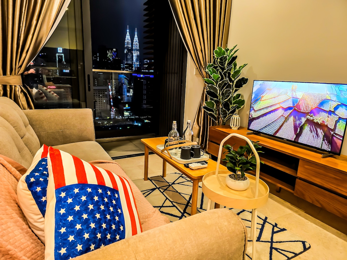 A stylish living room features a comfortable three-seater sofa with an accent pillow and a decorative plant. A coffee table is set with glassware. A large television displays vibrant colors, while floor-to-ceiling windows provide a view of the city skyline at night.
