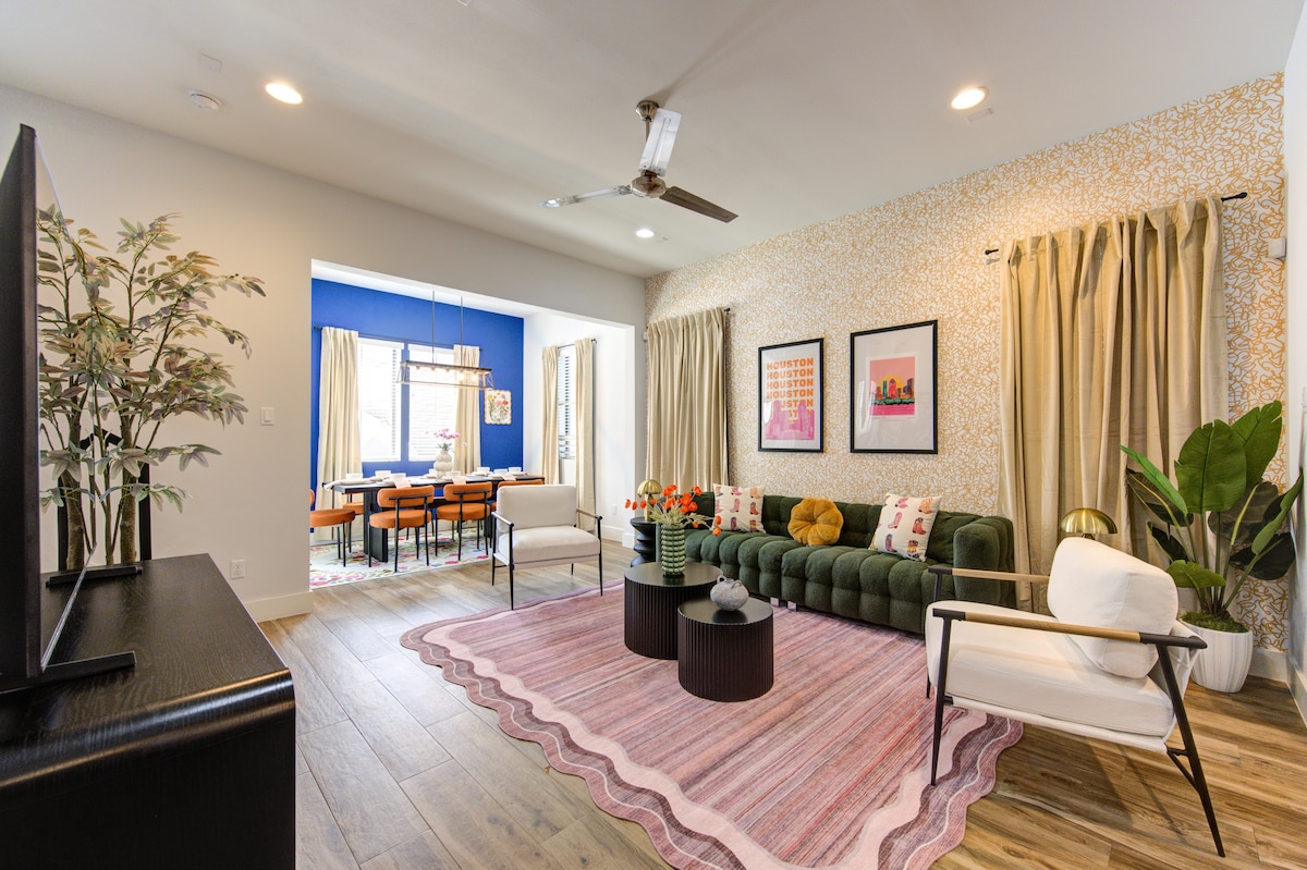 A spacious living room features a large green couch adorned with decorative pillows. A stylish area rug occupies the floor, complemented by a few small tables. Natural light pours in through bright curtains, with a view of the adjacent dining area highlighted by bold blue walls.