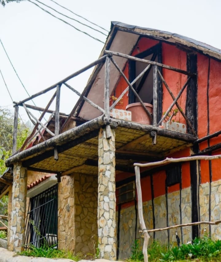 A charming cabin exterior is showcased with a combination of rustic stone and vibrant orange walls. A wooden balcony, supported by sturdy beams, offers a natural aesthetic. The surrounding area is framed by lush greenery, enhancing the connection with nature.