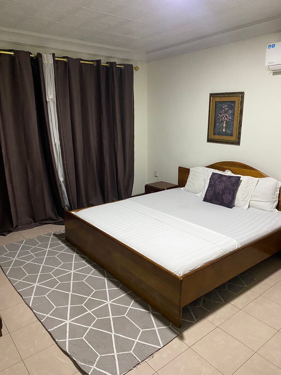 A modern bedroom features a wooden bed with a light-colored mattress, complemented by a decorative pillow. Dark curtains frame a window, while a geometric patterned rug lays on the tiled floor, enhancing the cozy atmosphere of the space.