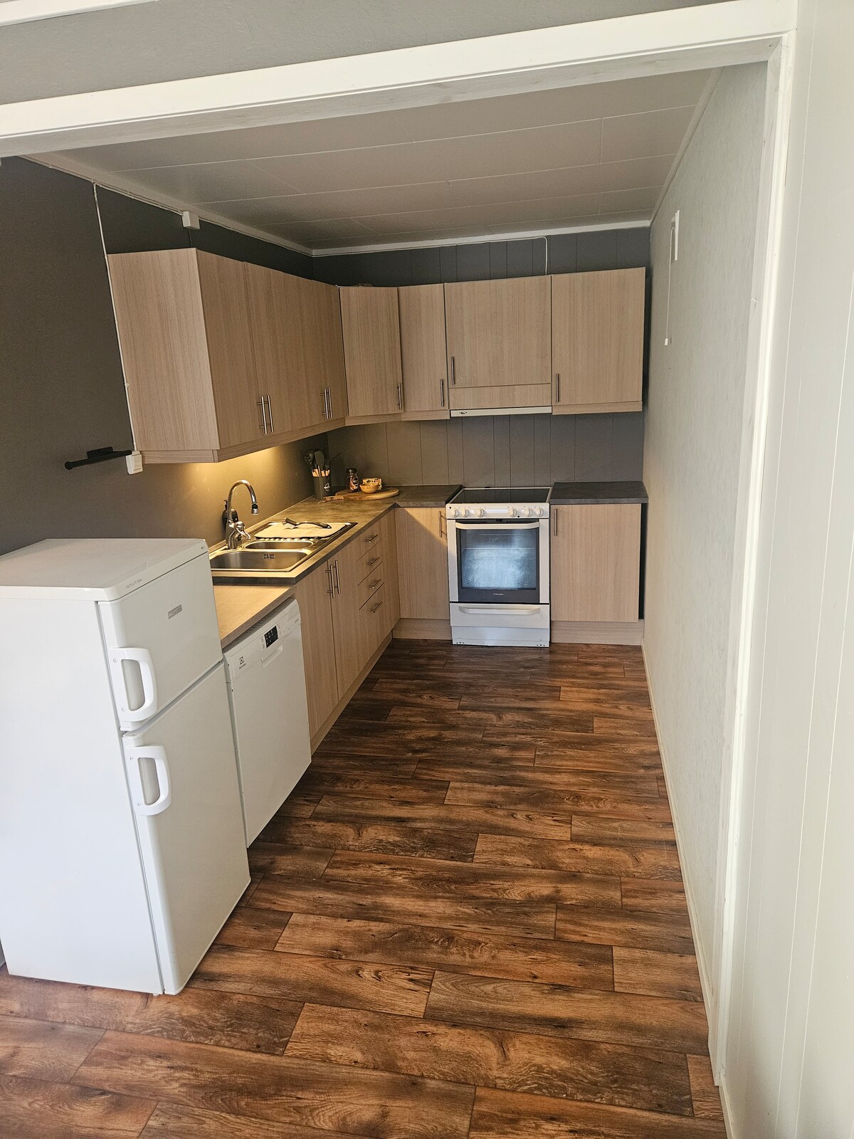 A functional kitchen features light wood cabinetry and modern appliances, including a refrigerator, dishwasher, and oven. Counter space is available for meal preparation, with additional items visible along the countertop. Natural light enters through a nearby window, enhancing the inviting environment.