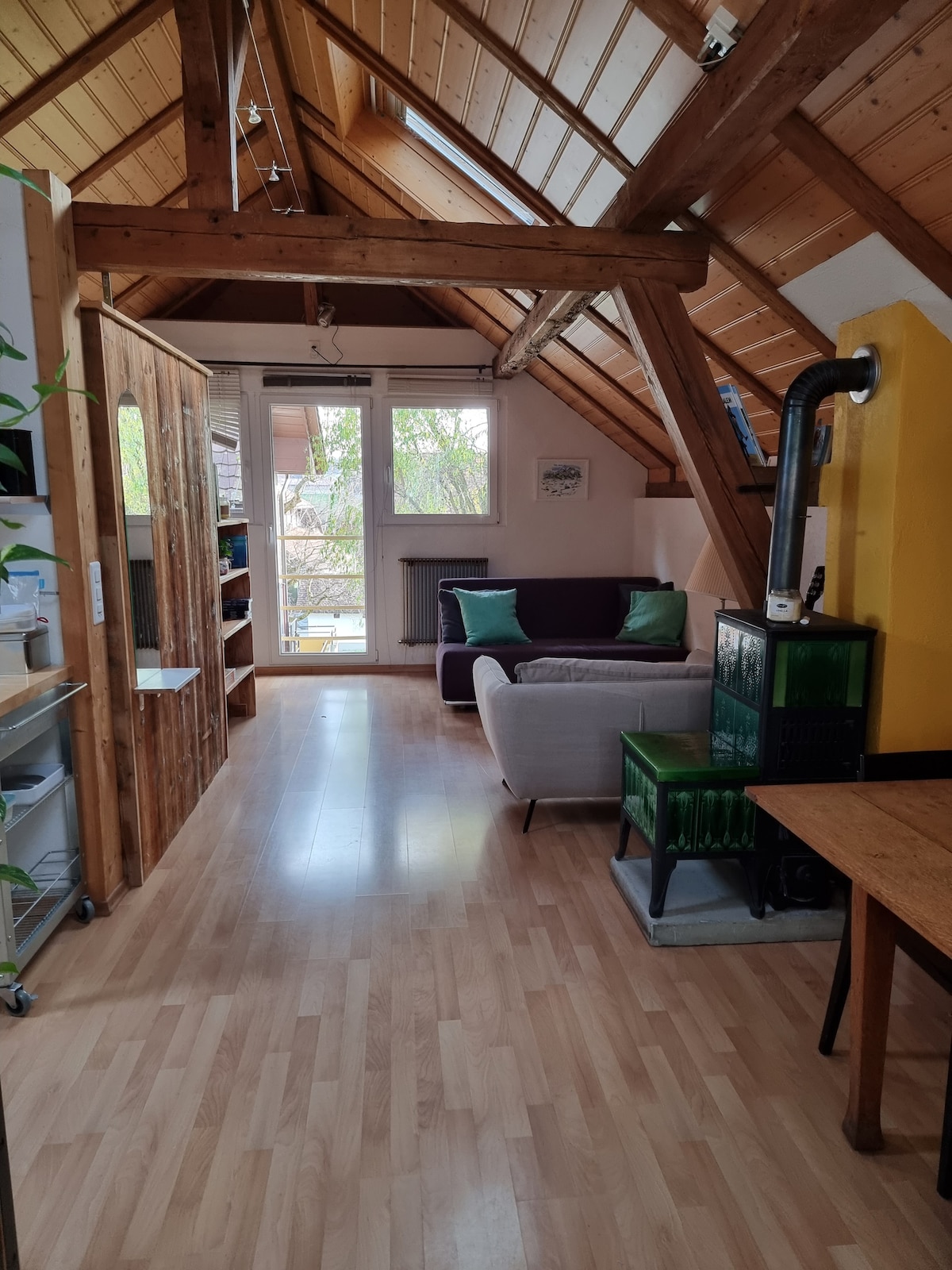 A lofted living area with sloped wooden ceilings features a mix of modern and rustic design elements. A light-colored sofa and a dark purple couch are complemented by green cushions. Large windows offer natural light, and a wooden table is positioned against the wall.