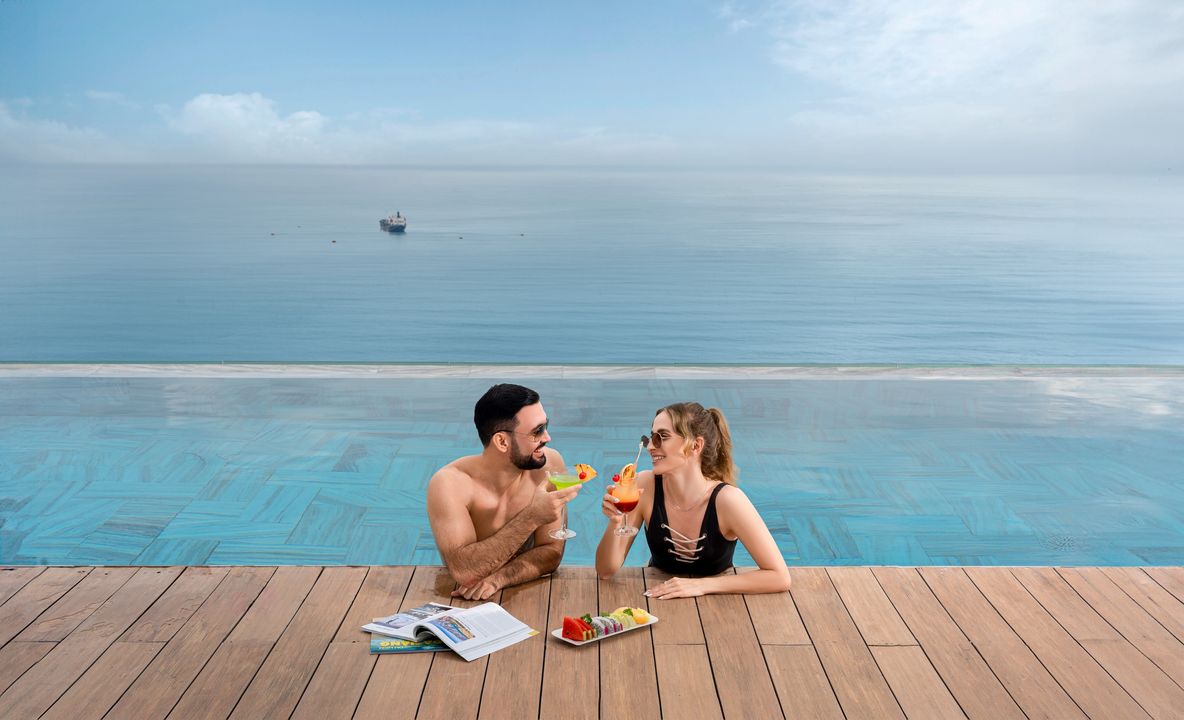 The infinity pool extends towards the horizon, offering a seamless view of the calm ocean. Two guests enjoy colorful beverages while relaxing on the pool's edge, surrounded by wooden decking. A tray of fresh fruit sits nearby, enhancing a serene atmosphere.