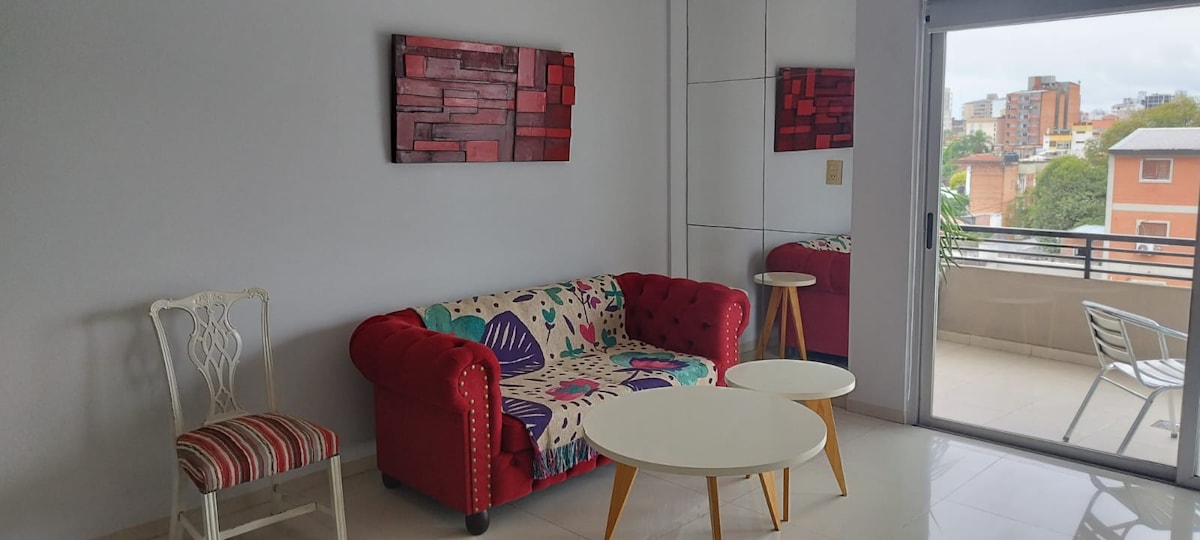 A bright living area features a colorful sofa with a patterned cover. A white round coffee table sits in front of the sofa, complemented by a light wooden side table. Artwork adorns the walls, while large windows offer natural light and views of the balcony.