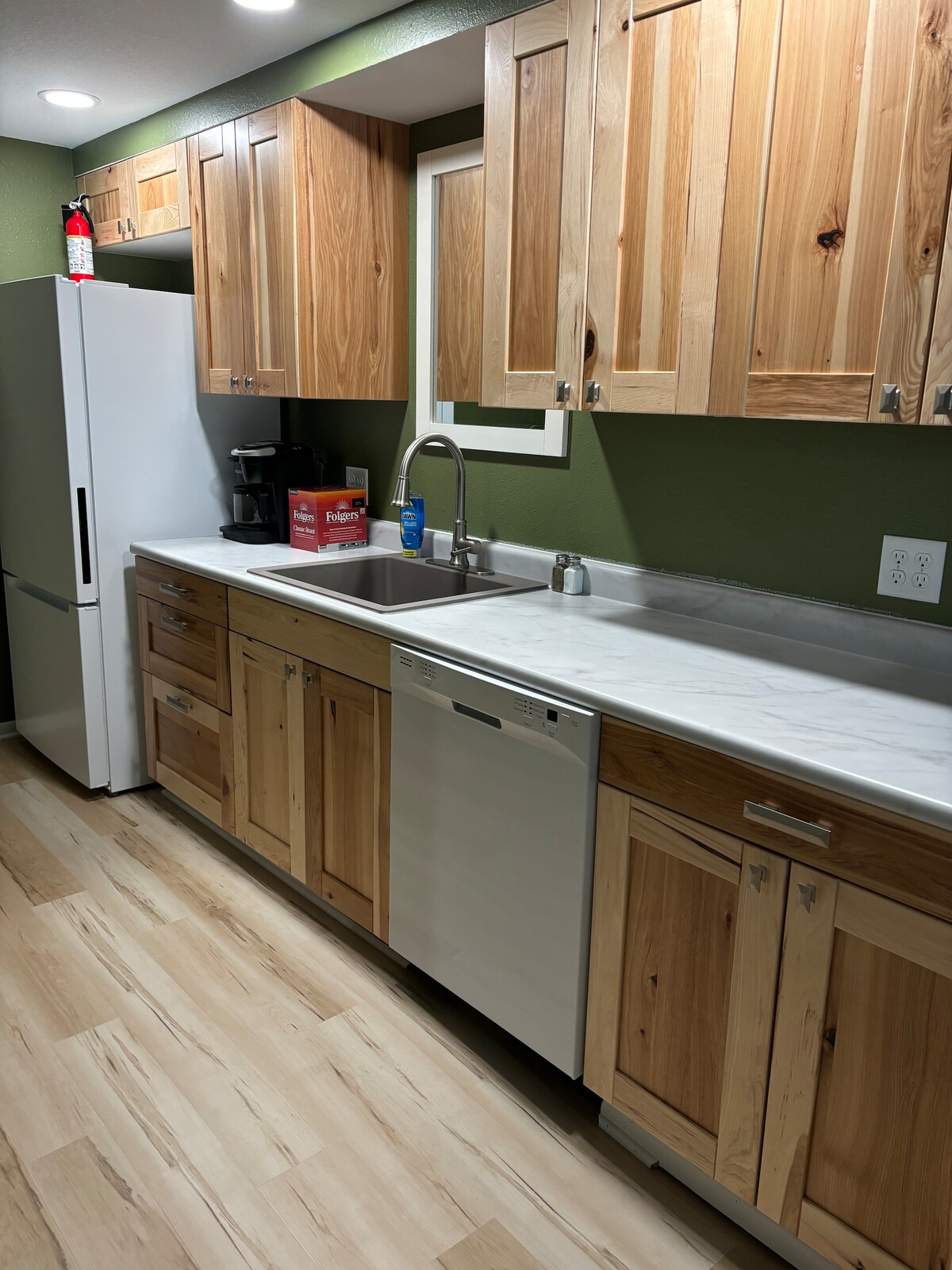 A modern kitchen features light wood cabinetry with a marble countertop. Essential appliances are included, such as a refrigerator, coffee maker, and dishwasher. Natural light is provided through a window above the sink, enhancing the clean and inviting space.