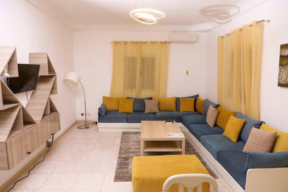 The living area is designed with a spacious L-shaped sofa featuring yellow and blue cushions. A low wooden coffee table is centrally positioned, while a mounted television is visible. Soft light fixtures hang above, and large windows are adorned with sheer yellow curtains.
