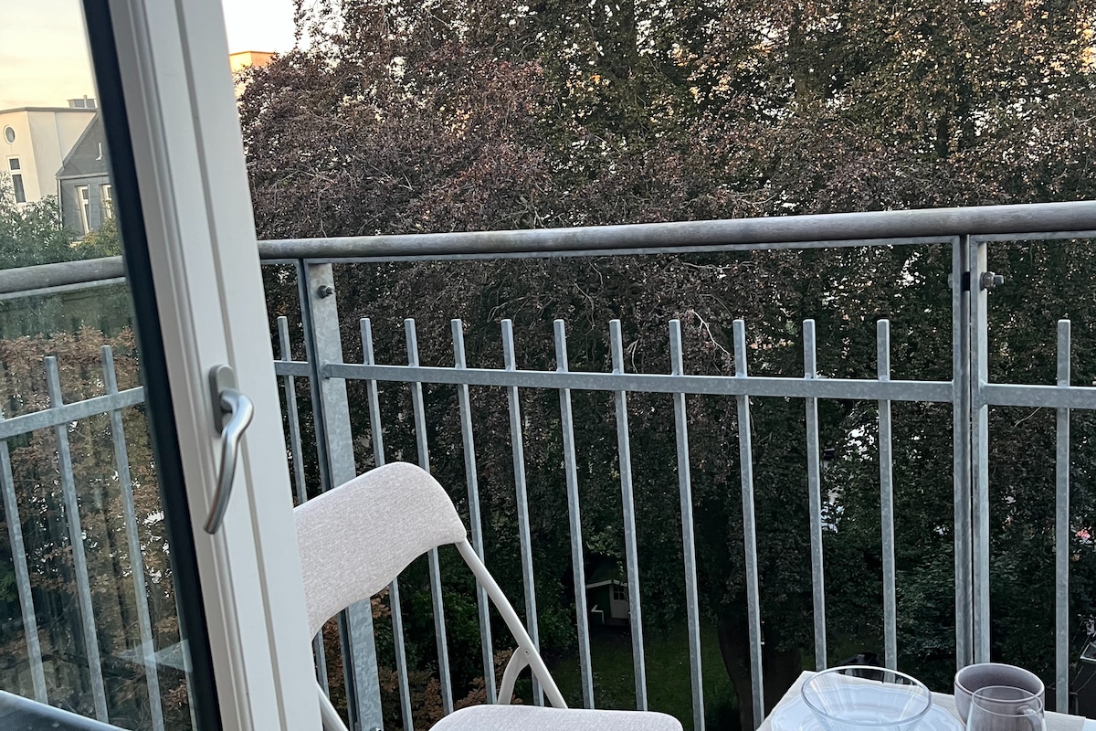 A cozy balcony area is featured, equipped with two light-colored chairs facing the greenery outside. A small table sits between the chairs, providing a spot for refreshments. The railing provides a clear view of the surrounding trees and garden below.