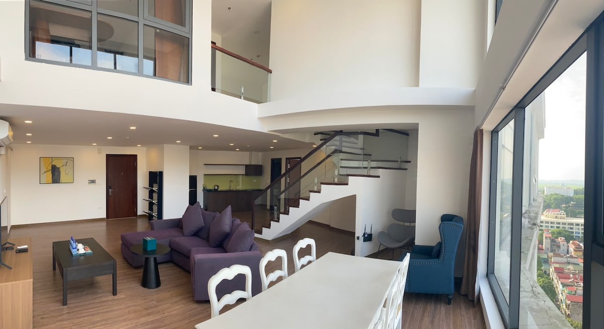 A spacious penthouse living area is shown, featuring high ceilings and large windows allowing for ample natural light. A comfortable purple sofa is arranged with a sleek coffee table, while a dining area with white chairs is visible. The open layout emphasizes a modern design.