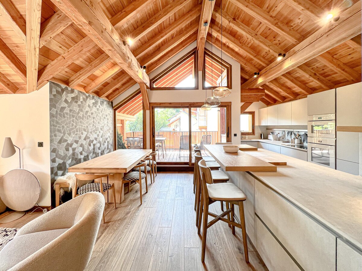 An open-plan kitchen and dining area features wooden beams and a stone accent wall. A large island with seating faces a dining table. Glass doors lead to a spacious terrace, allowing natural light to fill the room. Stylish pendant lights hang above the island.