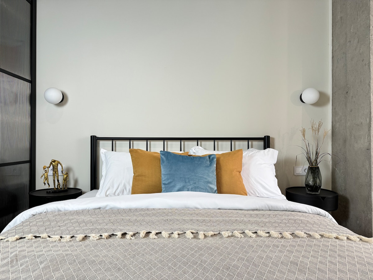 A well-presented sleeping area features a stylish bed with a neutral-colored blanket and an assortment of decorative pillows in shades of blue and gold. Modern bedside lamps and a small decorative arrangement add a touch of elegance to the space.