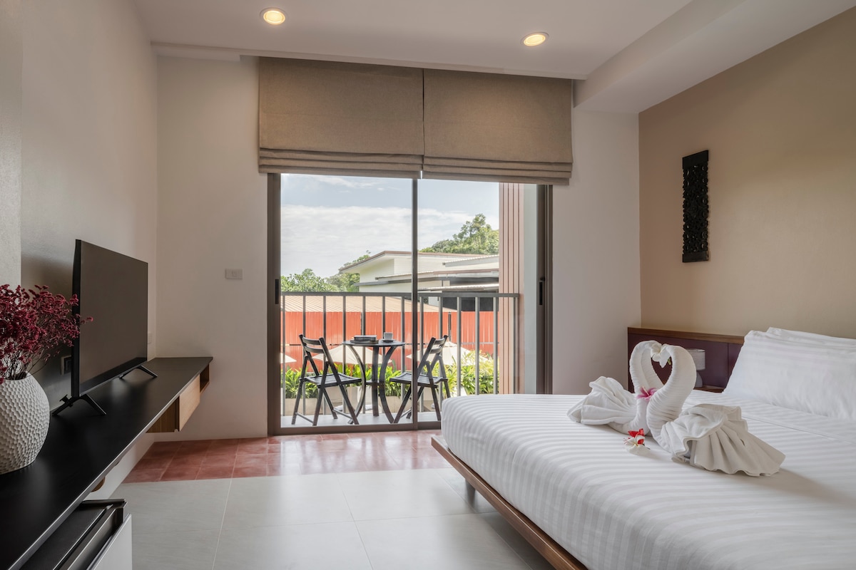 A well-lit room features a queen-size bed with crisp linens, a wall-mounted flat-screen TV, and a decorative arrangement of towels shaped into swans. A sliding glass door opens to a small balcony, providing views of the outdoor seating area and lush surroundings.