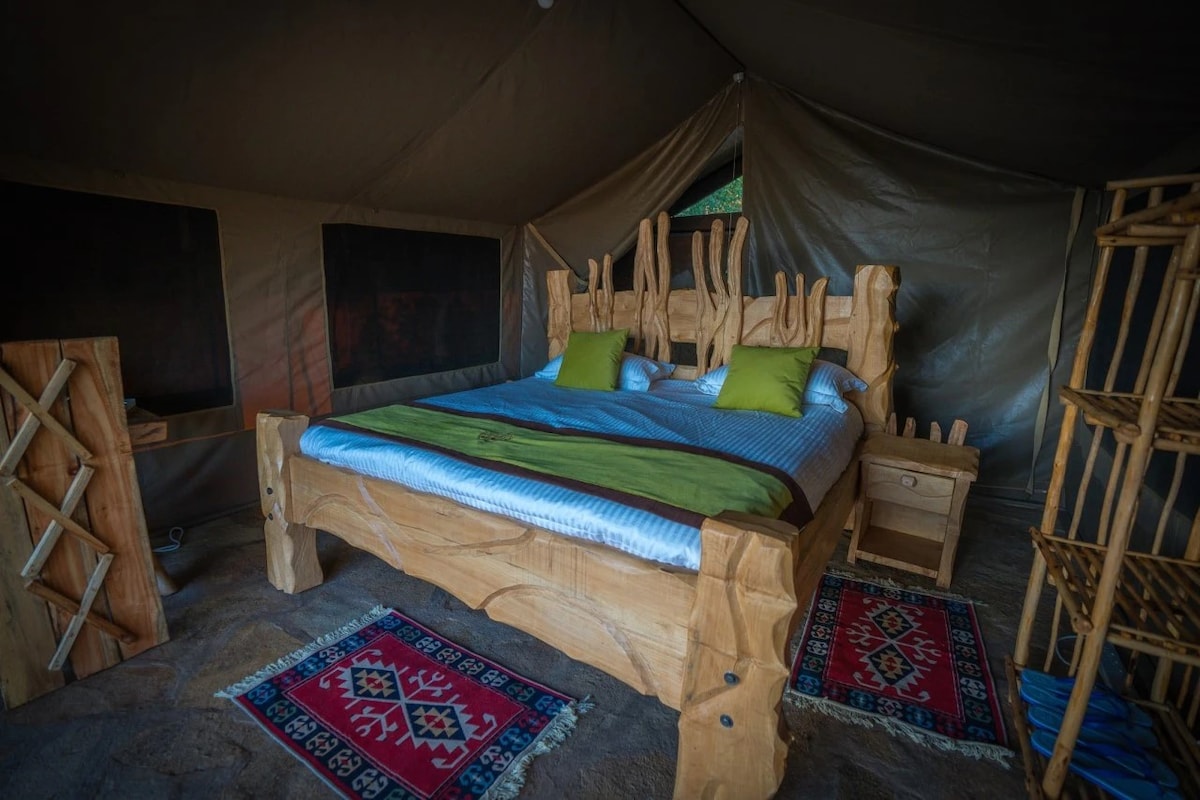 A spacious bed with a natural wood frame is centered in the tent, adorned with green accent pillows. Colorful rugs are positioned on the floor, providing warmth and comfort. Soft light filters through large windows, enhancing the inviting interior.