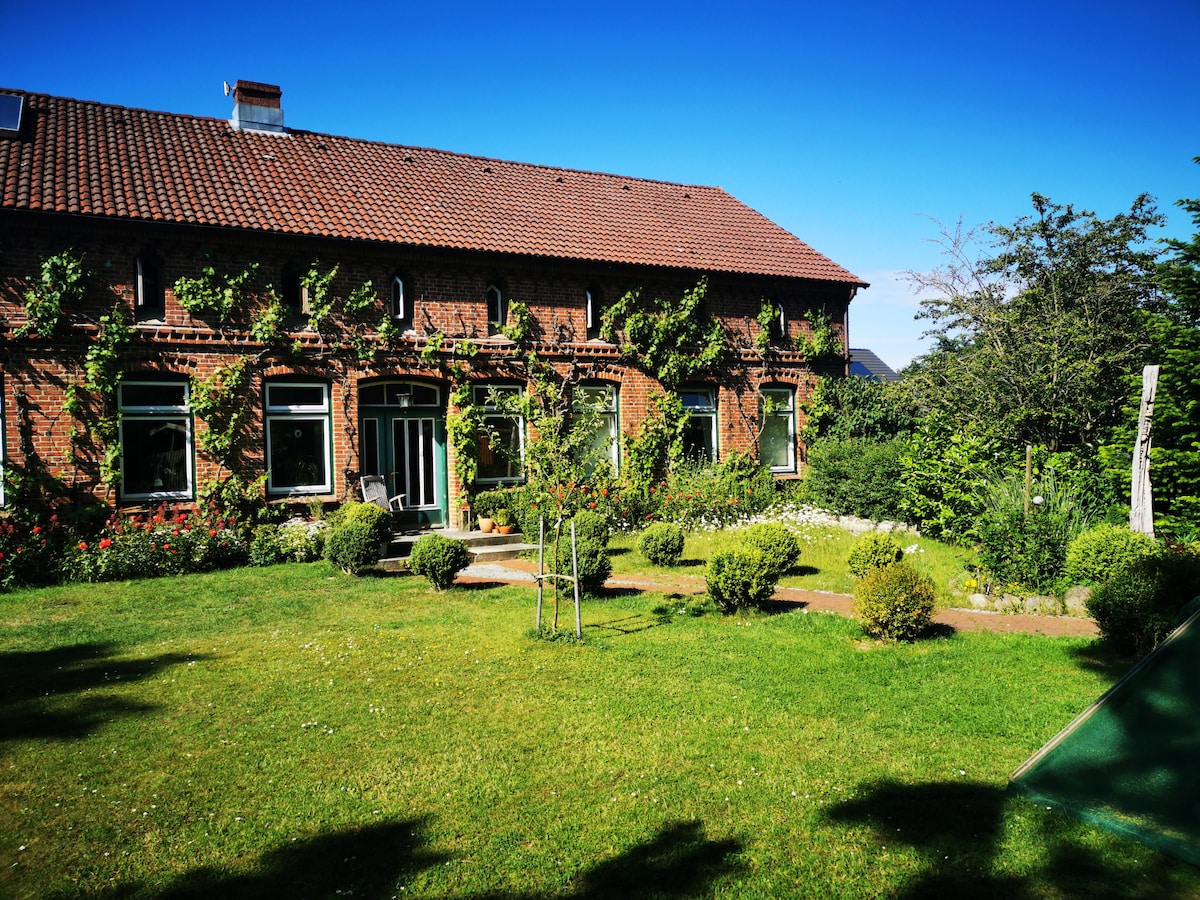 The exterior of a renovated farmhouse is shown, featuring a lush garden with well-maintained greenery. A brick façade is adorned with climbing plants, and several large windows invite natural light into the spaces inside. A seating area is visible on the patio.