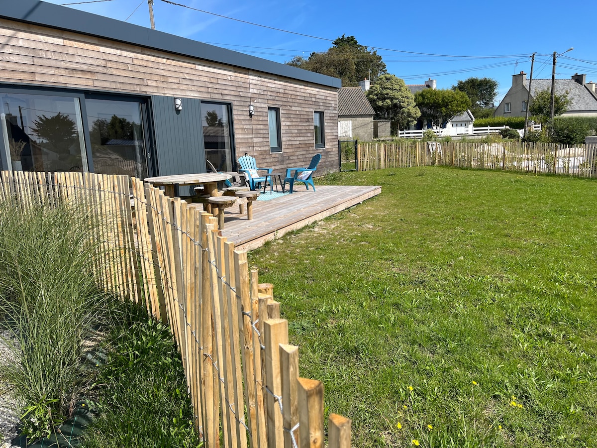 A well-maintained outdoor space is featured, with a wooden terrace hosting blue chairs and a table. A grassy area surrounds this space, and a bamboo fence encloses the property, providing privacy. The clear blue sky enhances the inviting atmosphere of the location.