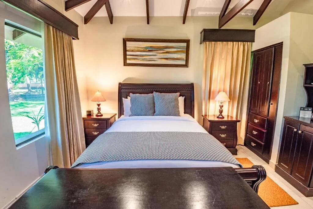 A cozy bedroom is presented with a spacious bed, dressed in light linens and accented with decorative pillows. Two nightstands with lamps flank the bed, while large windows allow natural light to fill the room. Rich wood tones are featured in the furniture and ceiling beams.