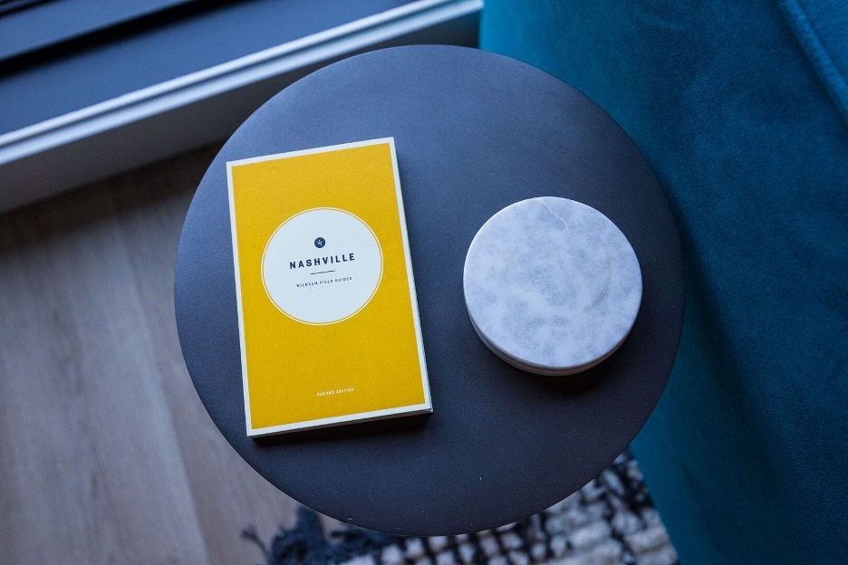 A small round side table is positioned next to a plush teal sofa. On the table, a vibrant yellow guidebook titled 'Nashville' is placed next to a sleek white marble coaster, providing a hint of local exploration.