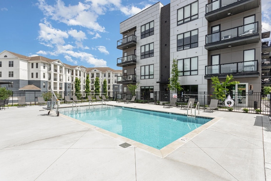 A bright pool area is surrounded by contemporary condominium buildings. The rectangular pool features a smooth, clear surface and several lounge chairs are positioned nearby. Green trees provide a touch of greenery against the modern architecture.