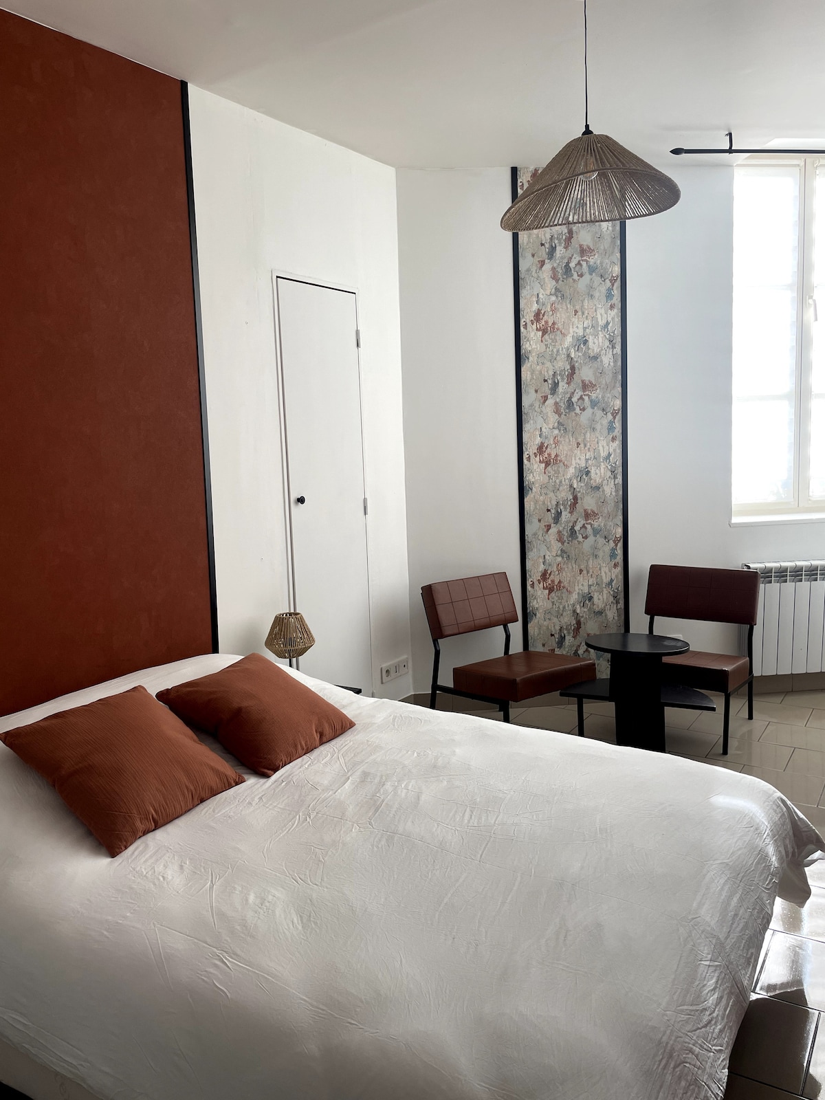 The sleeping area features a spacious bed with two decorative pillows. A small table and two chairs are positioned nearby, offering a cozy seating arrangement. A wall with floral patterns adds a subtle touch, while the bright window allows natural light to enter the room.