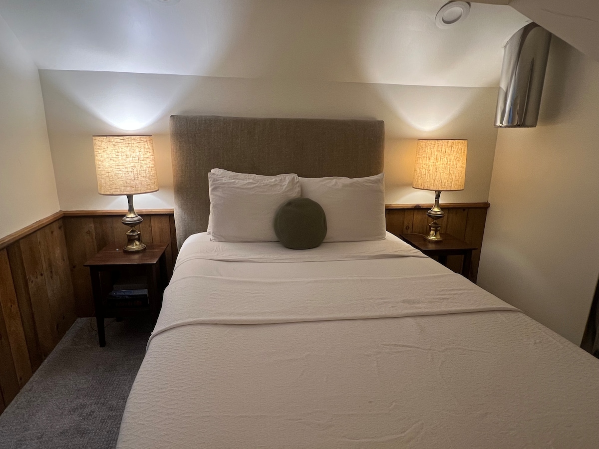 A well-appointed bedroom features a spacious bed with a solid headboard and neutral bedding. Two bedside lamps provide soft lighting, casting a warm glow. Wooden paneling complements the carpeted floor, creating a cozy atmosphere in the loft suite.