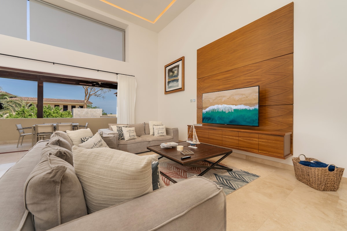 A spacious living area features a large sectional sofa arranged around a dark wood coffee table. A wall-mounted television is complemented by a wooden panel backdrop. Natural light enters through large windows, offering views of the outside. A dining table is visible in the background.