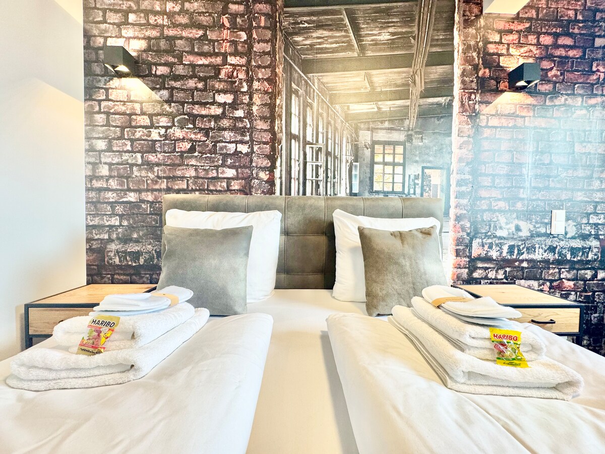 A cozy sleeping area features a stylish bed with soft linens. Neatly stacked towels are placed at the foot of the bed. The backdrop showcases a textured brick wall design, complemented by ambient wall sconces that provide gentle illumination.