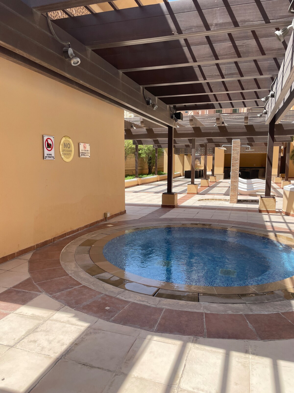 An outdoor area features a circular plunge pool surrounded by a tiled deck. Overhead, a wooden pergola provides shade, while sunlit tiled pathways lead to a landscaped garden area. Noticeable signage indicates pool regulations on nearby walls.