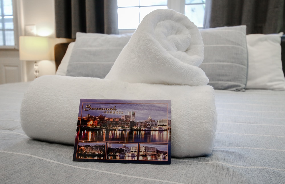A neatly arranged white towel is rolled and placed on top of a soft bedspread. A postcard featuring images of Savannah's waterfront is positioned beside the towel. Soft natural light filters through a nearby window, illuminating the inviting bedding.