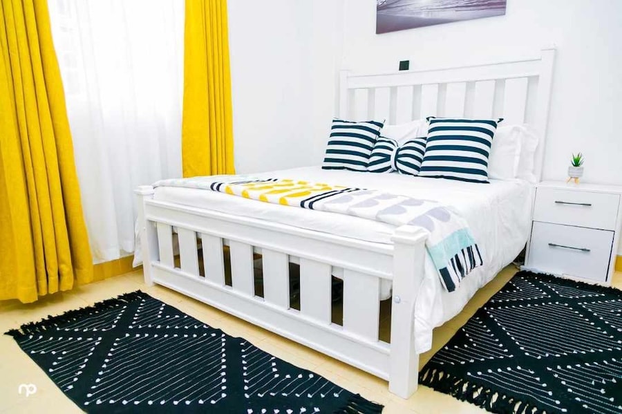 A bright sleeping area features a white wooden bed with a colorful blanket and striped pillows. Yellow curtains frame a window, allowing natural light to illuminate the space. Two decorative black rugs add texture to the floor, enhancing the room's contemporary style.
