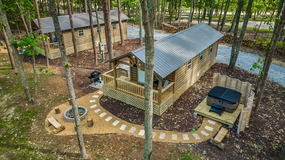 A rustic cabin is surrounded by trees in a serene woodland setting. A gravel path leads to the entrance, while a private hot tub and fire pit are positioned on a wooden deck. Outdoor seating and a grill are visible, creating a welcoming atmosphere.