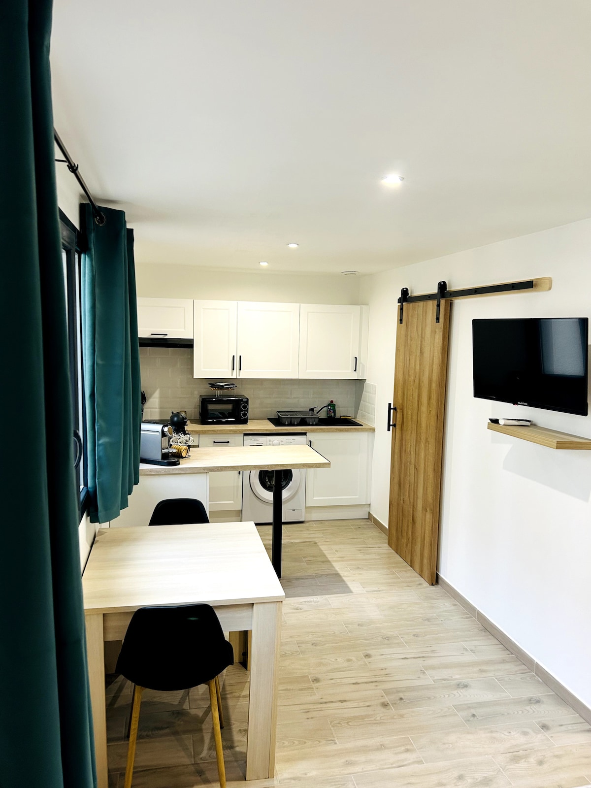 A compact kitchen and dining area are visible, featuring sleek white cabinetry, a countertop with modern appliances, and a small dining table. A wall-mounted TV can be seen nearby, along with a sliding barn door that adds a functional touch to the space.