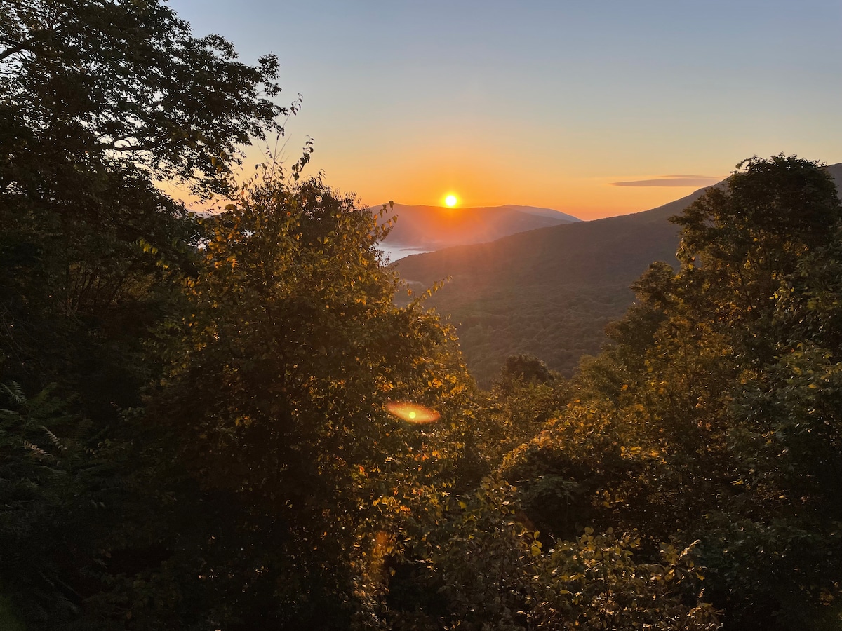 A sunset is seen from the cabin's surroundings, casting warm golden hues over the distant mountains. Lush greenery frames the view with trees and foliage, creating a serene atmosphere. The horizon showcases a soft transition of colors, blending the sky with the landscape below.