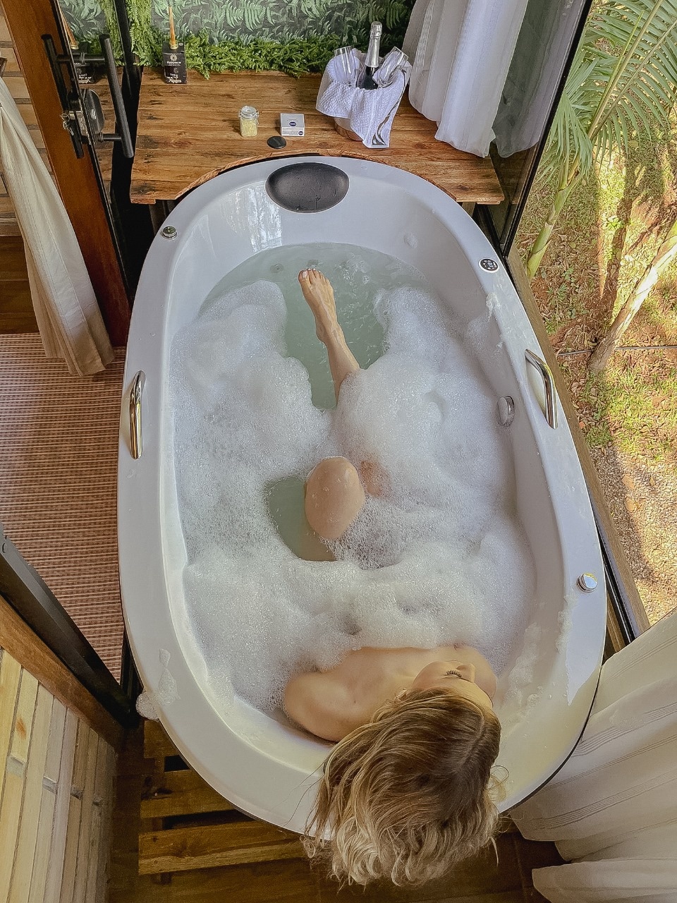 A freestanding soaking tub sits invitingly among the greenery, filled with bubbles. Natural light filters through large windows, creating a serene atmosphere. Wooden surfaces complement the modern design, while a bottle and glass are placed nearby, hinting at a moment of relaxation.
