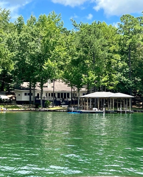 The lakehouse is surrounded by trees, showcasing a blend of natural greenery and modern design. A spacious deck extends along the side of the house, leading to a large covered dock. The clear water reflects the house and trees, creating a serene lakeside setting.