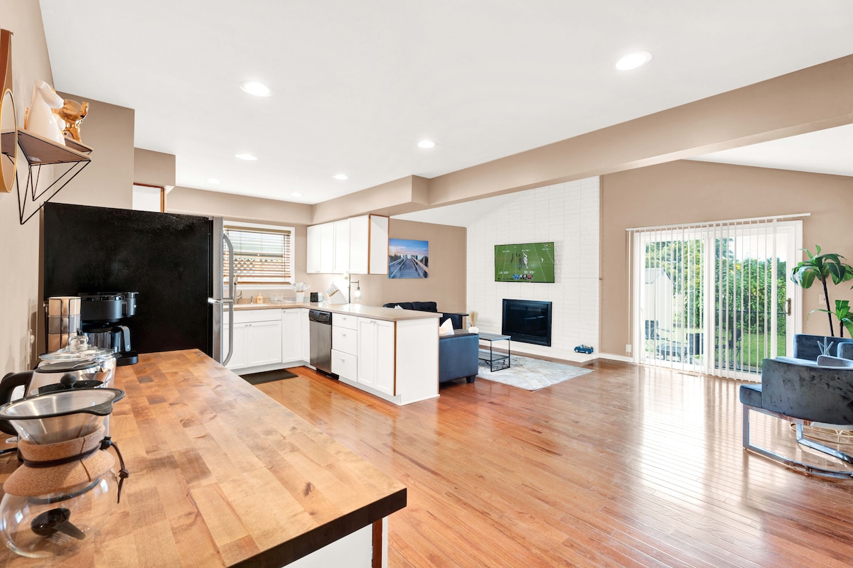 The spacious living area features an open kitchen with modern appliances and a large wooden countertop. A comfortable seating arrangement is present, with a view of the outdoor area through sliding glass doors. Natural light fills the space, enhancing the bright atmosphere.