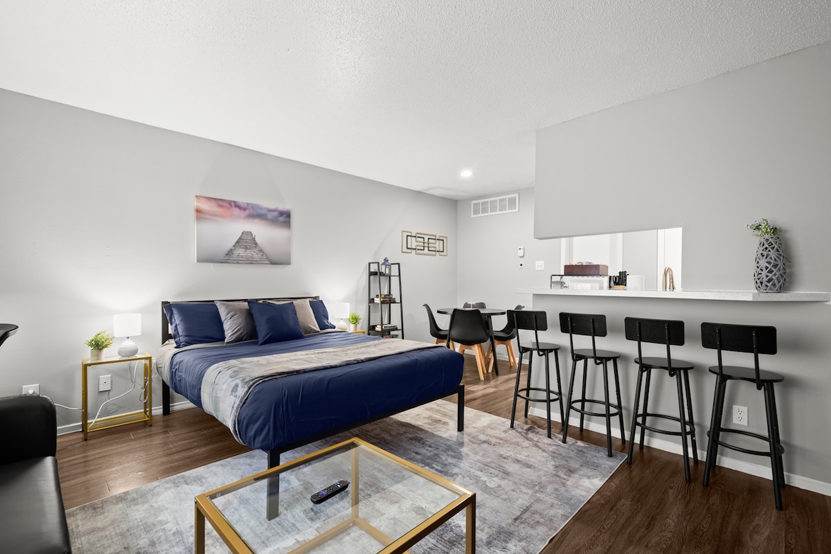 A spacious studio layout showcases a cozy queen bed with premium linens. A stylish futon adds seating, while a dining table with stools invites shared meals. Natural light fills the room, highlighting a modern aesthetic and inviting ambiance.