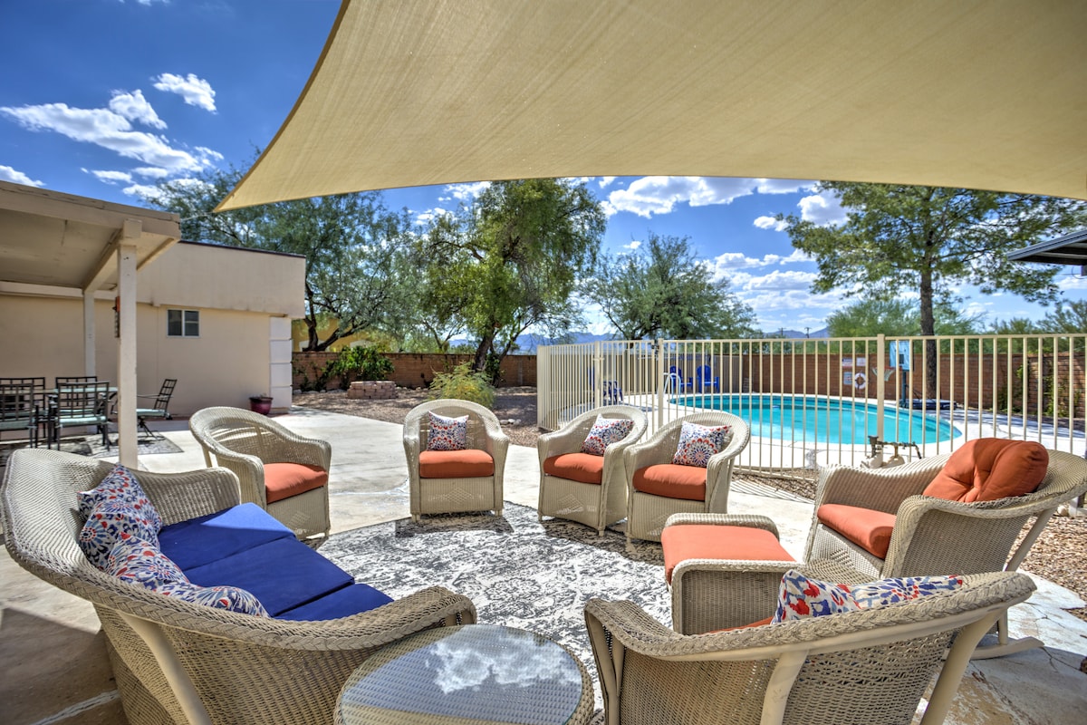 An outdoor seating area features several plush chairs arranged around a central circular table, with a large shaded canopy overhead. In the background, a sparkling pool reflects the blue sky, surrounded by lush greenery and a fence providing privacy.