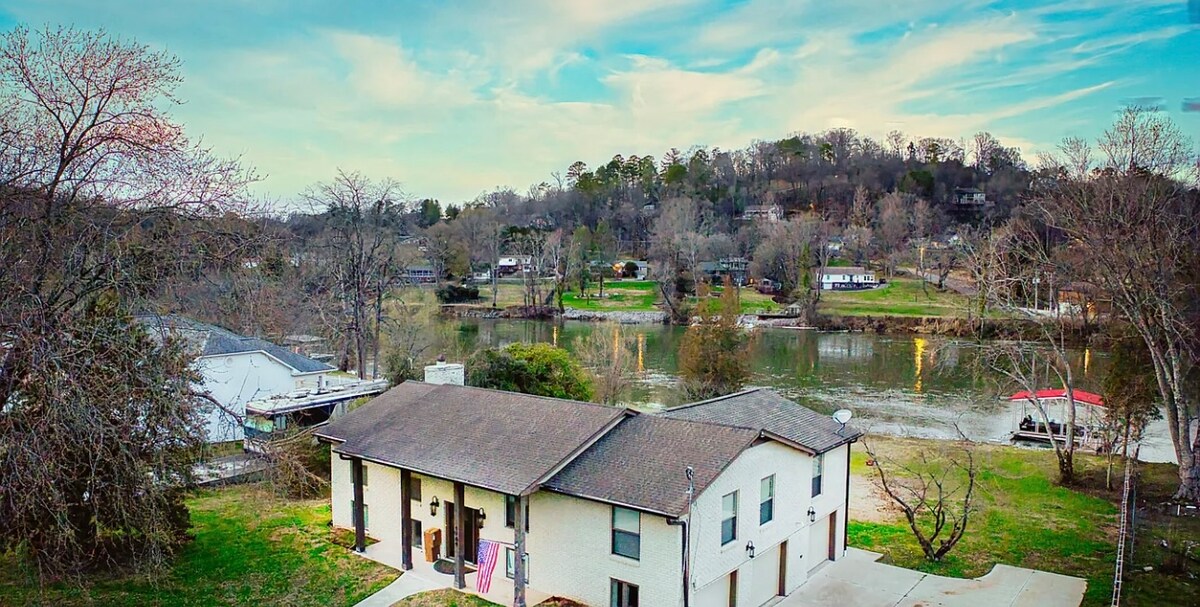 An aerial view showcases the riverfront home, nestled among trees and overlooking the water. The two-story structure features a neutral exterior, with a spacious driveway in front and a serene landscape of lush greenery in the background.