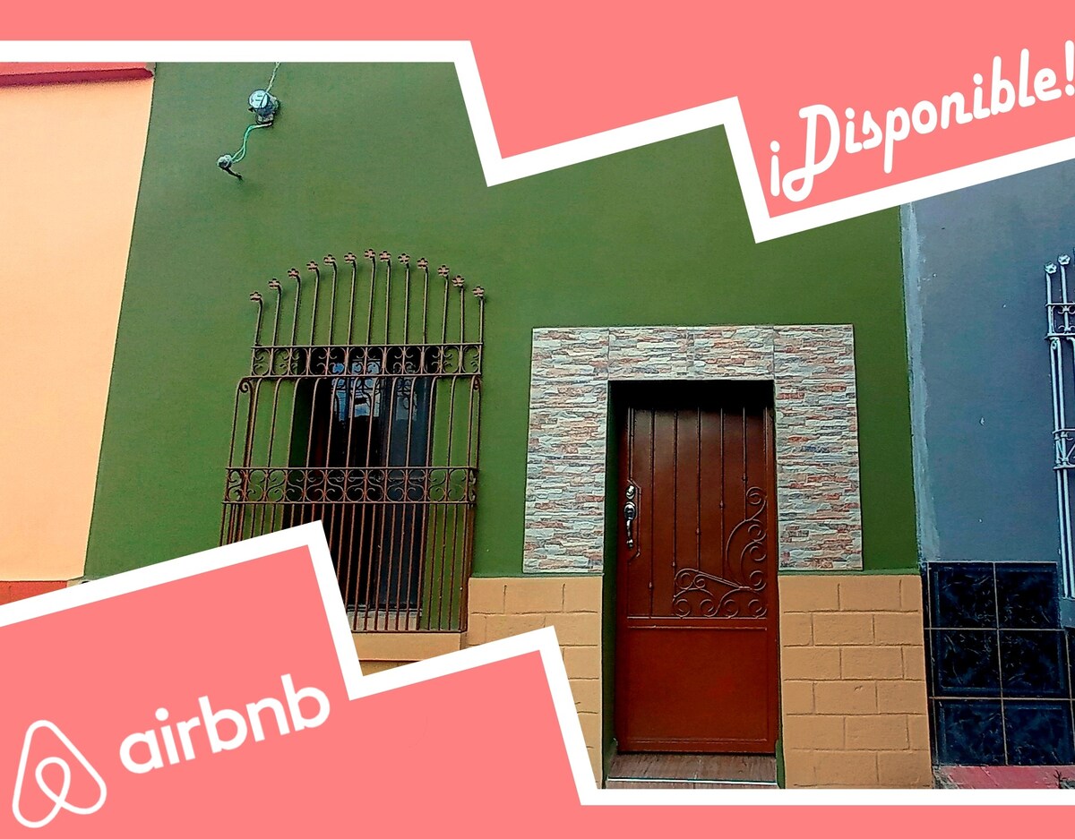 A vibrant green exterior features a decorative stone border surrounding the main entrance door. The door is wooden with an intricate design, flanked by a wrought-iron window. The textured walls exhibit a combination of warm earth tones, presenting an inviting street presence.