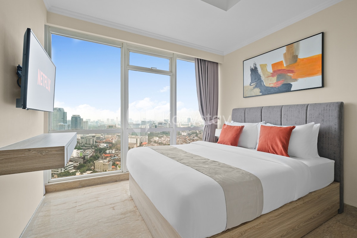 A bright master bedroom features a spacious bed with a light-colored bedspread and decorative pillows. A large window offers expansive city views, complemented by a mounted television displaying the Netflix logo. Light curtains frame the window, allowing natural light to fill the room.