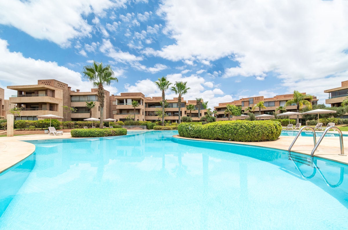 A serene outdoor pool is surrounded by lush greenery and palm trees, reflecting the clear blue sky above. Surrounding structures feature modern designs, and several lounge chairs are positioned near the water's edge for relaxation.