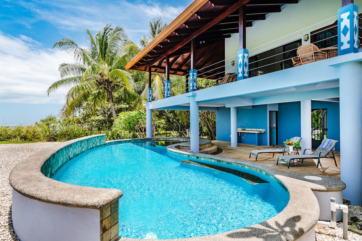 A saltwater swimming pool is bordered by a spacious patio area featuring lounging chairs and tropical landscaping. The modern architecture includes a covered porch with decorative columns, showcasing an inviting blue and white color scheme under a clear sky.