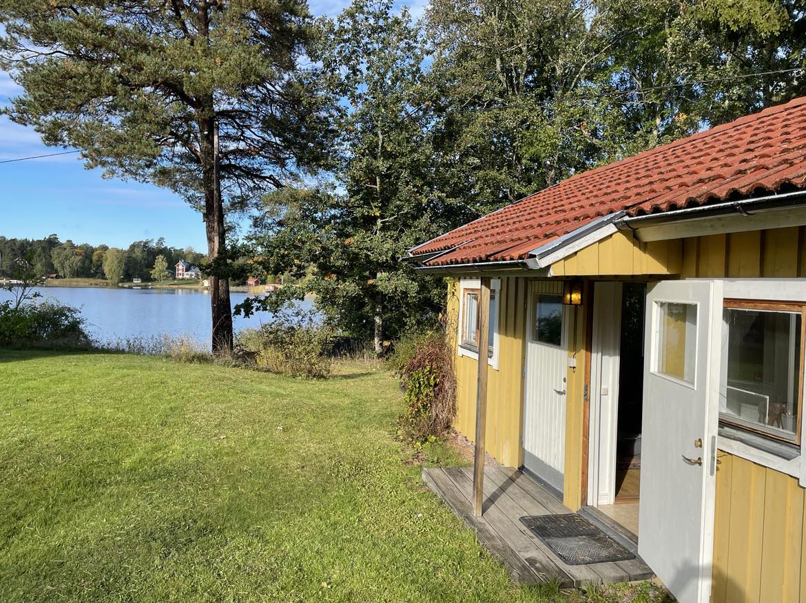 A quaint cottage exterior is visible, featuring yellow walls and a red-tiled roof. Two white doors open onto a wooden deck, leading to a lush green lawn. The serene lake can be seen in the background, framed by trees under a clear blue sky.