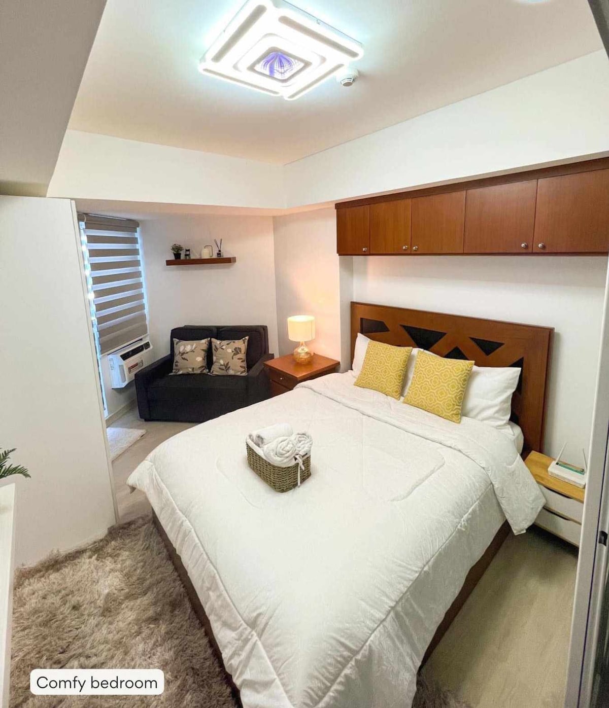 A cozy bedroom is showcased, featuring a spacious bed with light bedding and accent pillows in yellow. A small lamp illuminates a bedside table, while a comfortable seating area is positioned nearby. Natural light filters through partially open window blinds.