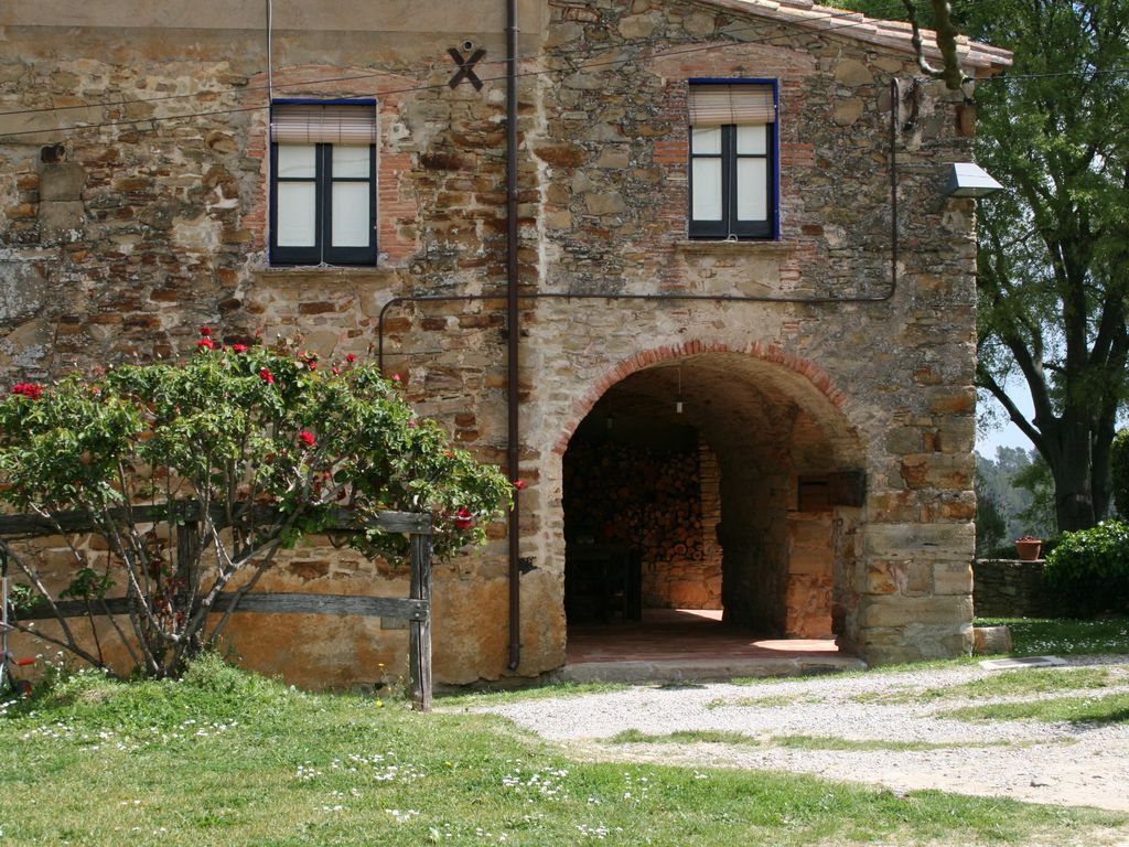 The sturdy stone exterior of the house showcases a blend of natural materials, with archways leading to an inviting entry. A flowering bush adds color to the entrance, while a stack of firewood is visible within the arched area. The well-maintained grassy area enhances the overall rustic appeal.