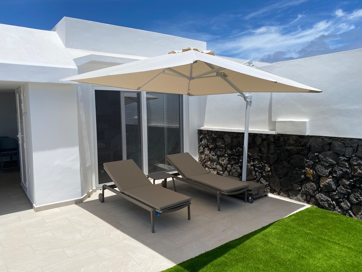 A terrace area is featured with two sun loungers positioned beneath a large umbrella, providing shade. The surrounding area includes well-maintained stone walls and artificial grass, enhancing the outdoor space for relaxation. Natural light illuminates the terrace, creating an inviting atmosphere.