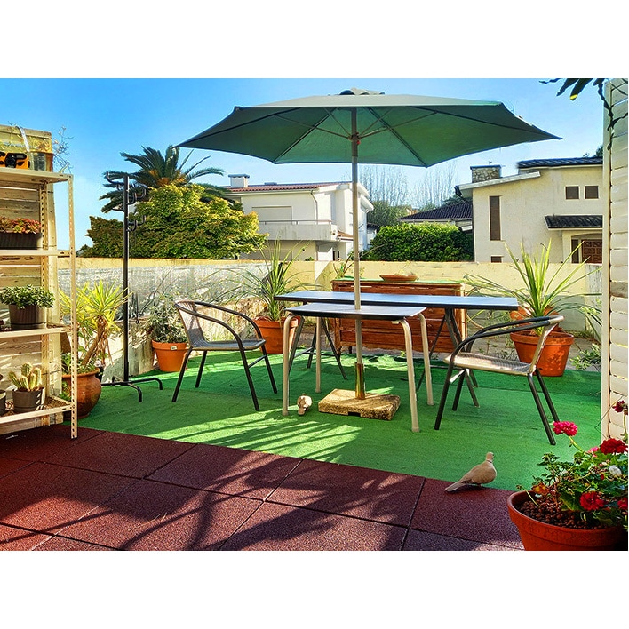 A terrace area is presented, featuring a table with an umbrella surrounded by four chairs. Potted plants add greenery, while a small stone is seen on the ground. The space is illuminated by natural light, enhancing the inviting setting.
