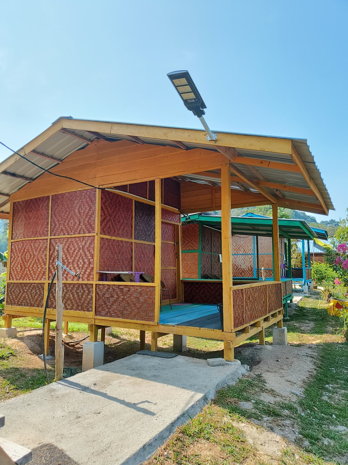 A traditional wooden structure features colorful woven walls and a sloped roof. The building is elevated on concrete pillars with a small porch area. Surrounding greenery and vibrant flowers enhance the natural setting.